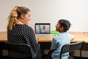 Adult sitting next to child at a desk with a laptop that shows a Speech Therapist on a video call.