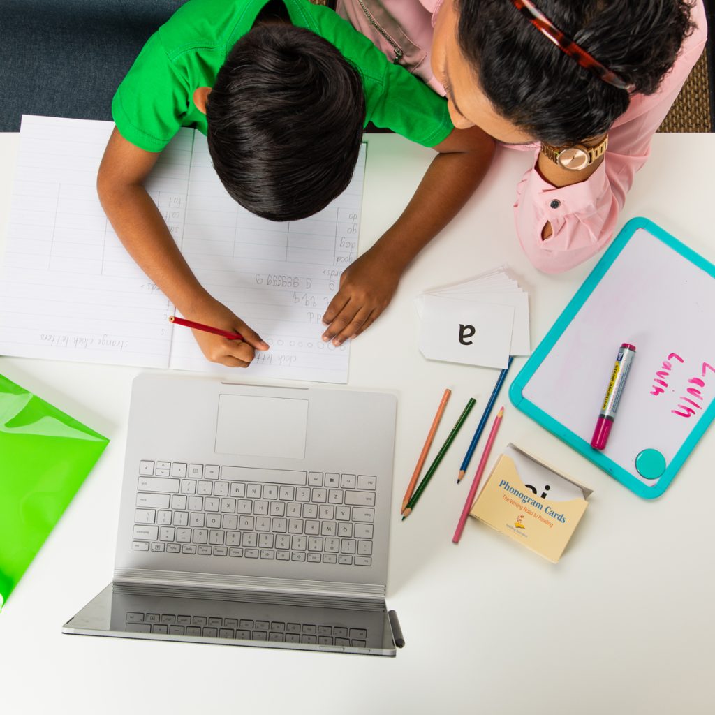 Square image of birds eye view of a child practicing writing at a desk with an adult sitting next to them